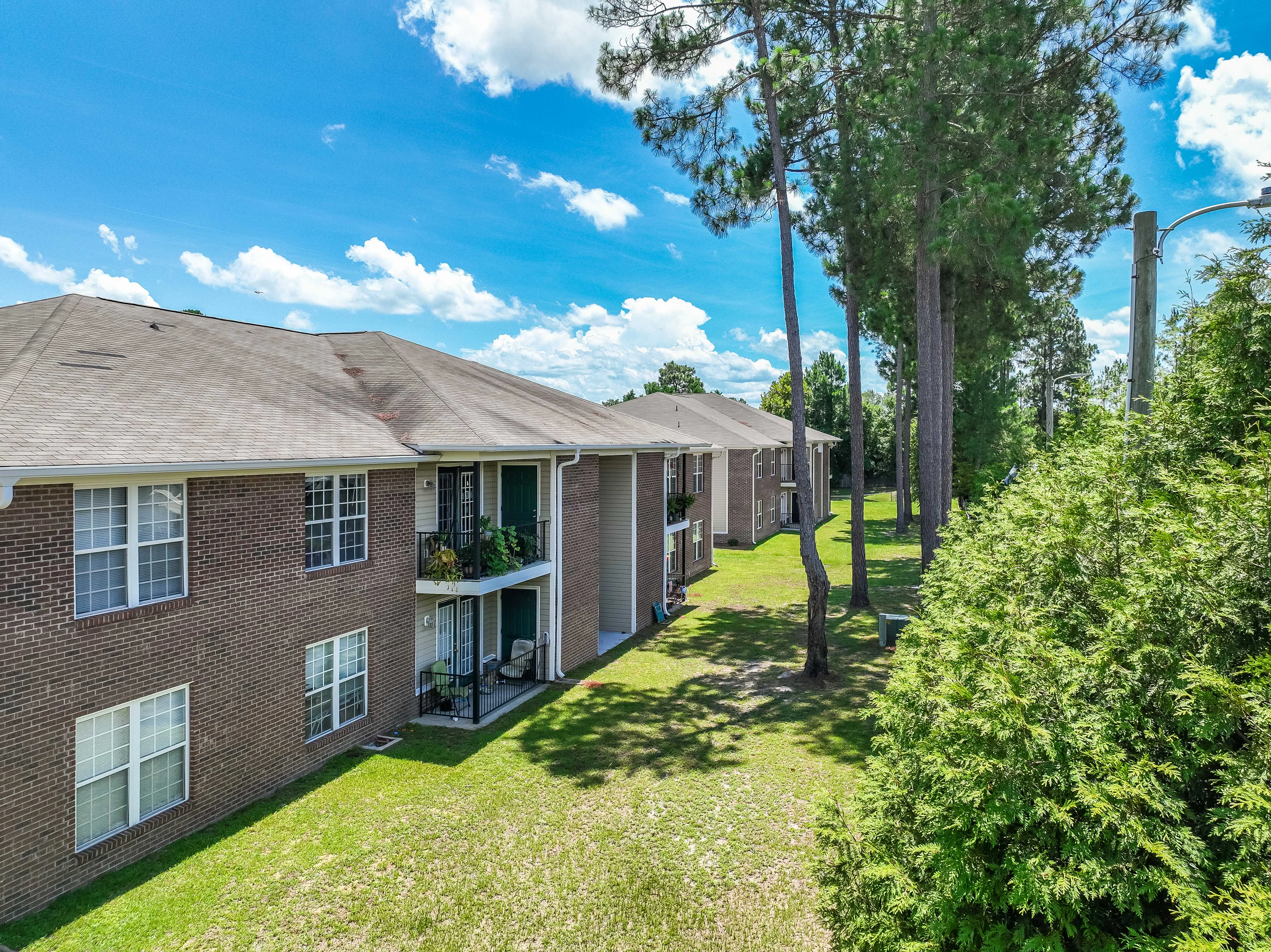 Exterior view of a brick apartment complex surrounded by greenery and trees under a blue sky with clouds.