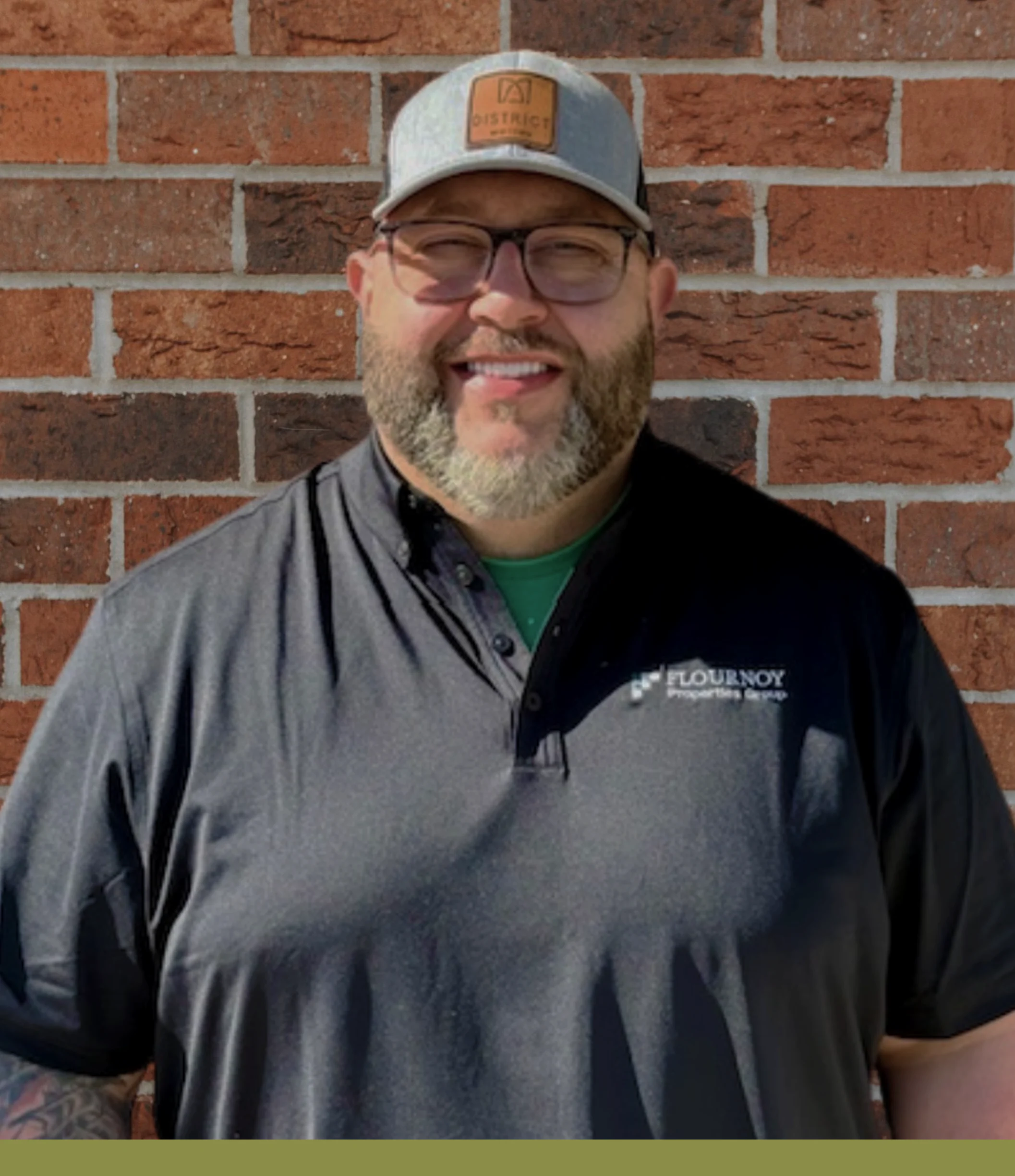 Outdoor Portrait of a Man A smiling man with a beard wearing a baseball cap and a dark polo shirt with a logo, standing against a brick wall.