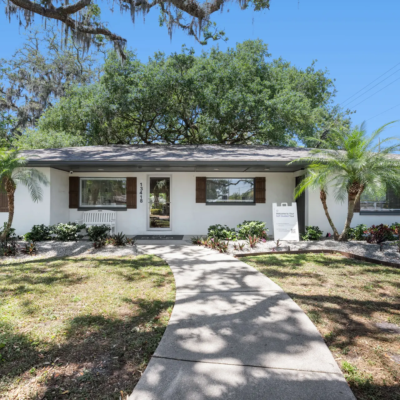 A front view of a single-story building surrounded by greenery and landscaping. The building has a welcoming walkway leading to the entrance, and a sign is visible on the right side.