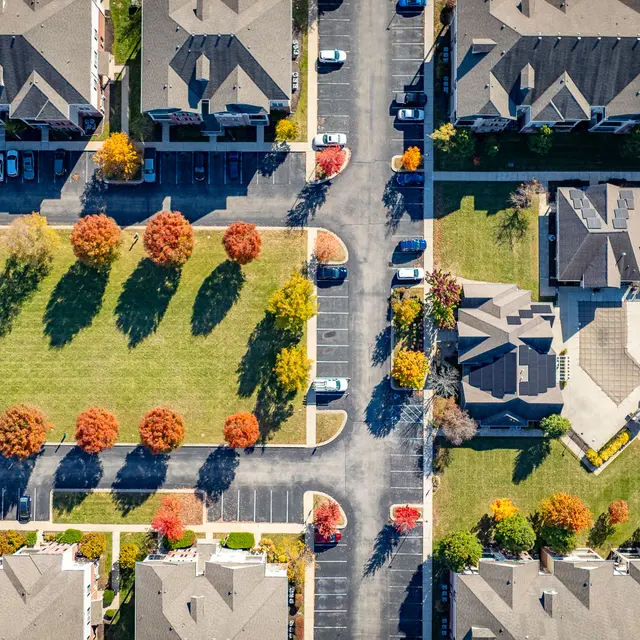 Aerial view of an apartment complex featuring multiple buildings arranged around green spaces and parking areas, with trees showing autumn foliage.