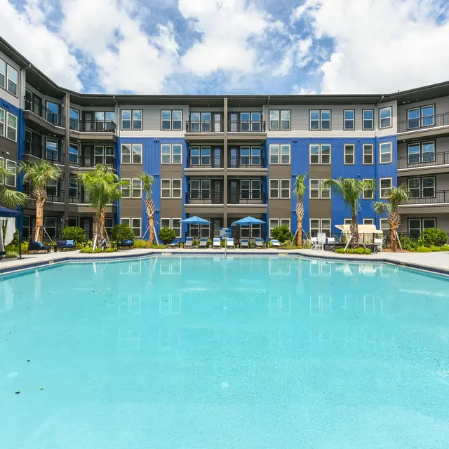 Stunning Pool Area of a Modern Apartment Community A modern apartment complex featuring a blue exterior, surrounding a clear swimming pool with palm trees and lounge chairs.