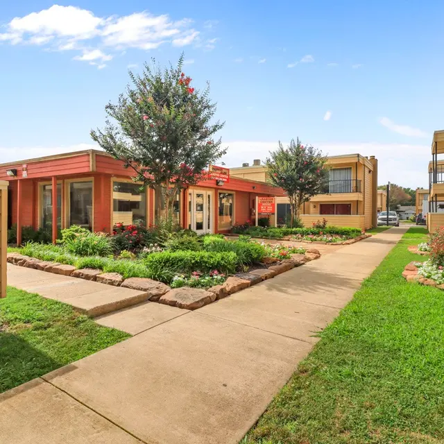 A landscaped apartment complex featuring a pathway leading through green grass and flower beds with small shrubs and trees. There are several buildings with a warm color palette in the background.