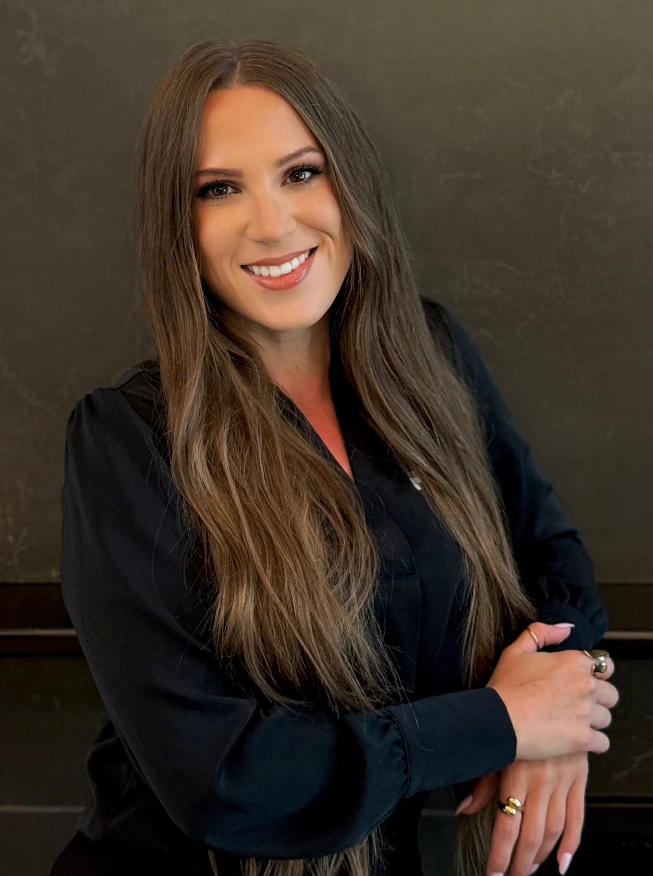 A woman with long brown hair poses in front of a dark background, wearing a black blouse with a friendly smile, showcasing a confident demeanor.