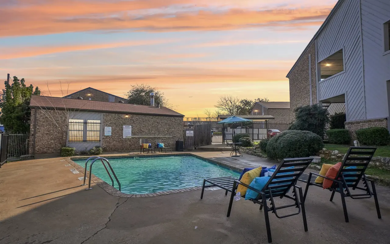A welcoming pool area at sunset, featuring a clear pool, colorful cushions on lounge chairs, and a cloudy sky with pastel hues.