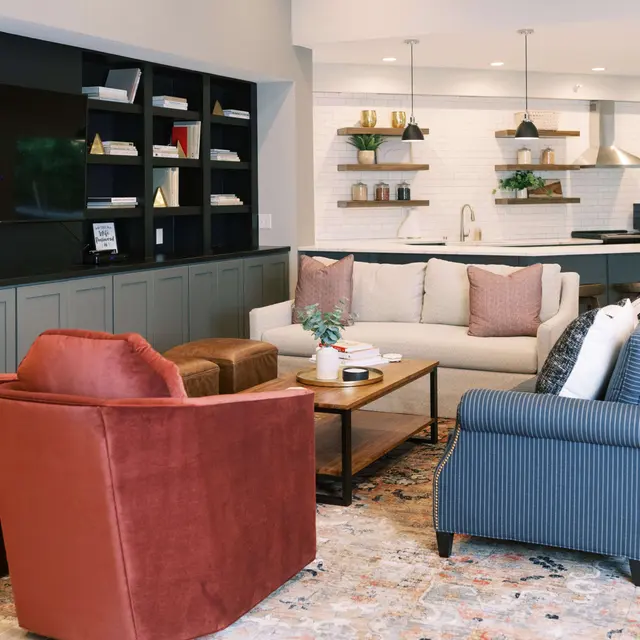 A stylish living room featuring a sectional sofa with decorative pillows, two armchairs in red fabric, a wooden coffee table with a small plant centerpiece, a large flat-screen TV on the wall, and a sleek kitchen area in the background with minimalistic decor.