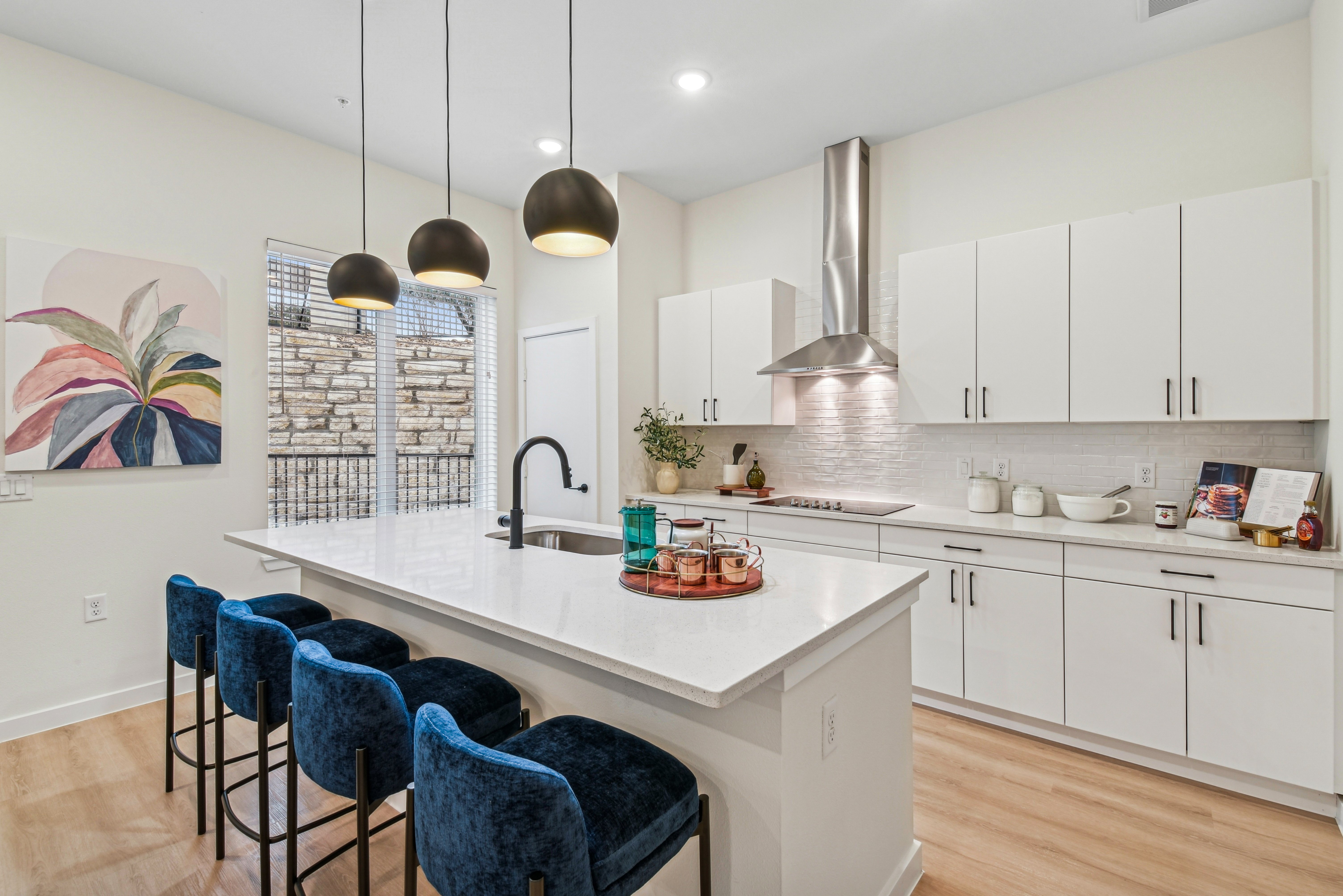 A modern kitchen featuring white cabinets, an island with bar stools, and pendant lighting. The decor includes a colorful wall art and a stone wall visible through the window.