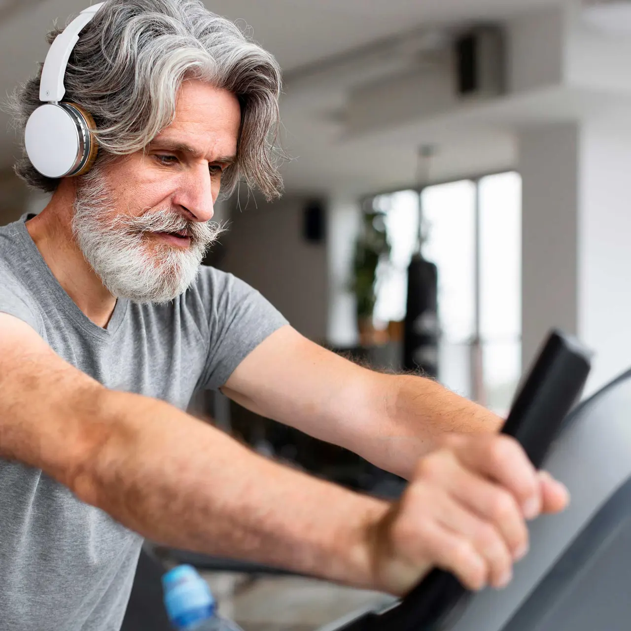 Elderly Man Exercising at the Gym An elderly man with gray hair and a beard using an elliptical trainer in a gym, wearing white headphones. He is focused on his exercise.