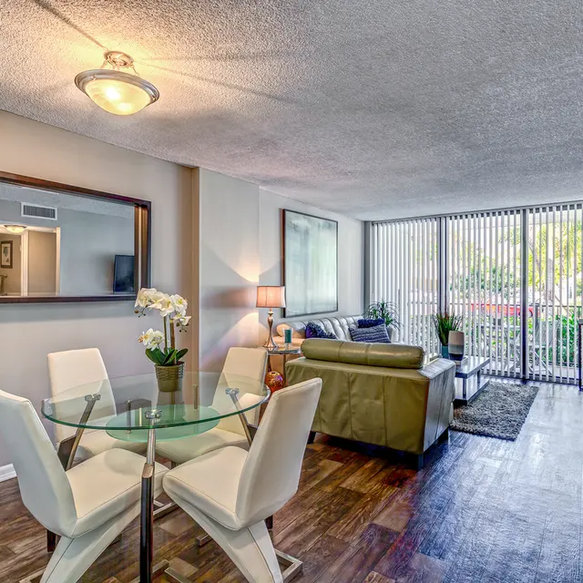 A modern living room featuring a glass dining table with white chairs, a green couch, and a large window with vertical blinds letting in natural light. A stylish mirror hangs on the wall, and plants enhance the decor.