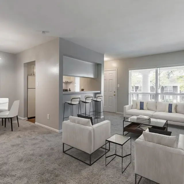 A stylish and modern living room space with a neutral color palette. The area features a cozy white sofa with decorative cushions, two accent chairs, and a coffee table. A dining area with a round table and four chairs is visible on the left side, along with a light fixture. The room is well-lit by natural light coming through large windows.