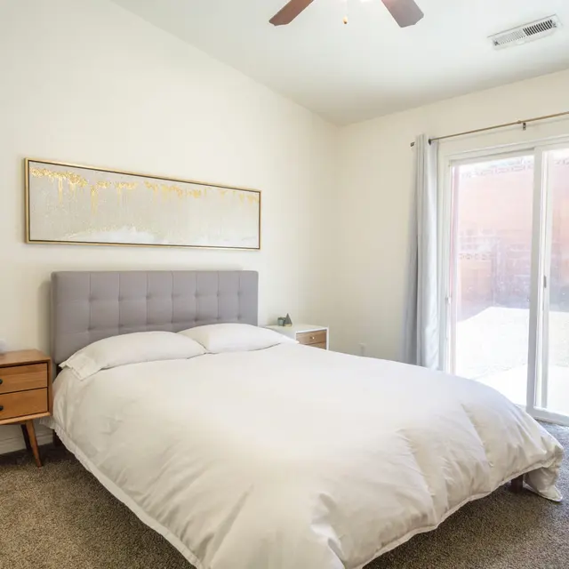 A bright and minimalist bedroom featuring a king-sized bed with a light gray headboard, a wooden nightstand beside it, and sliding glass doors leading to an outdoor area.