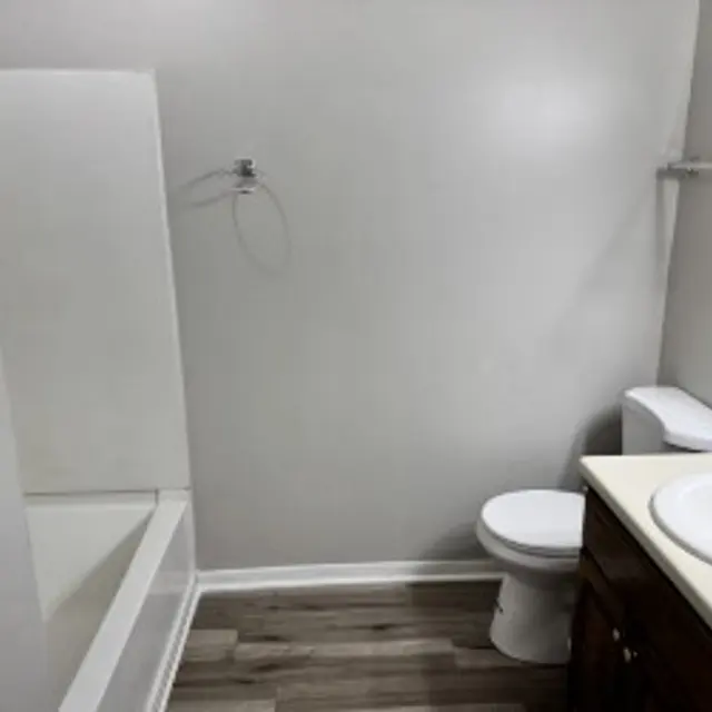 A clean and simple bathroom featuring a white bathtub, a white toilet, and a vanity with a sink. The walls are painted light gray, and the floor is covered with light wood-like laminate. There is a towel rack and minimal decorations, creating an open and uncluttered space.