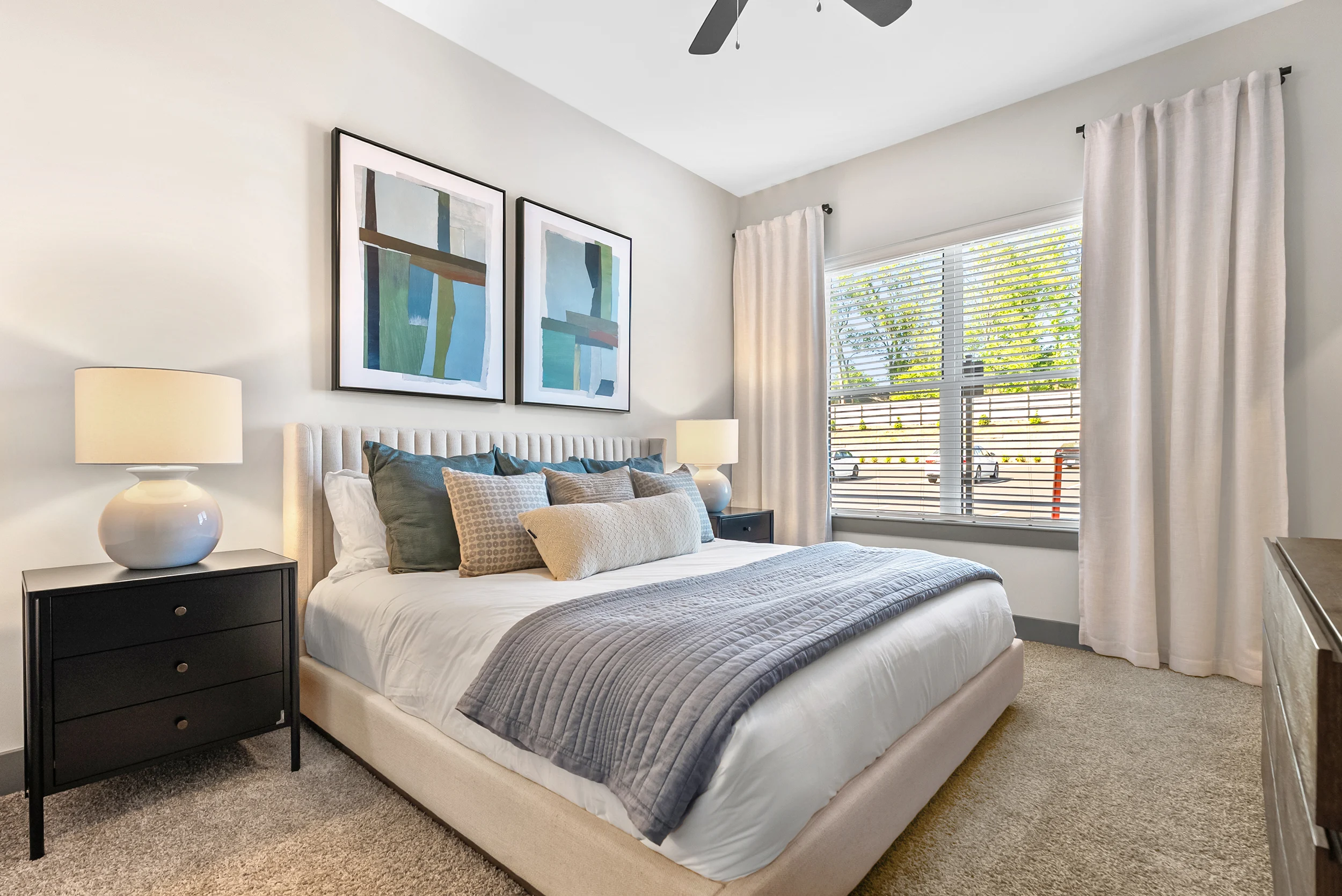 A modern bedroom featuring a queen-sized bed with decorative pillows, a bedside table with a lamp, and a large window with light curtains. There are two abstract framed artworks on the wall.