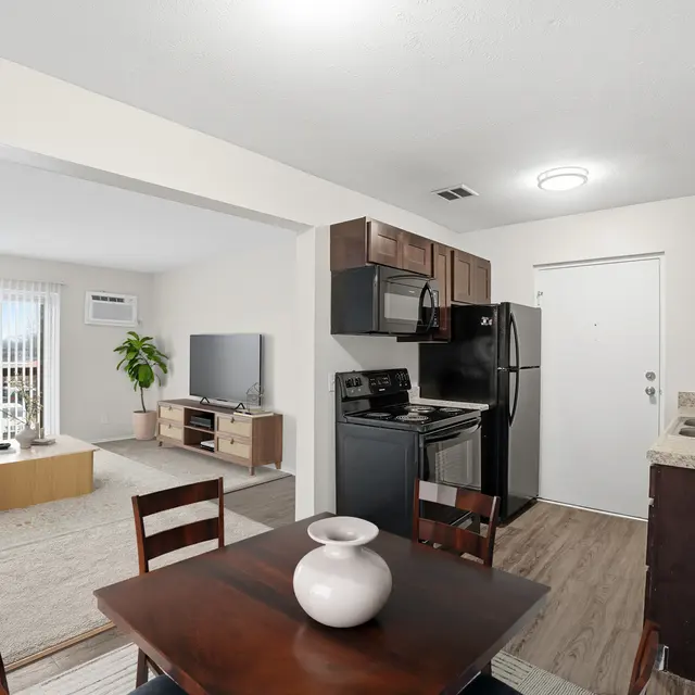 A modern apartment interior featuring a kitchen and living area. The kitchen has dark cabinetry and stainless steel appliances, while the living area is furnished with a couch, coffee table, and a television. There is natural light coming through large windows.