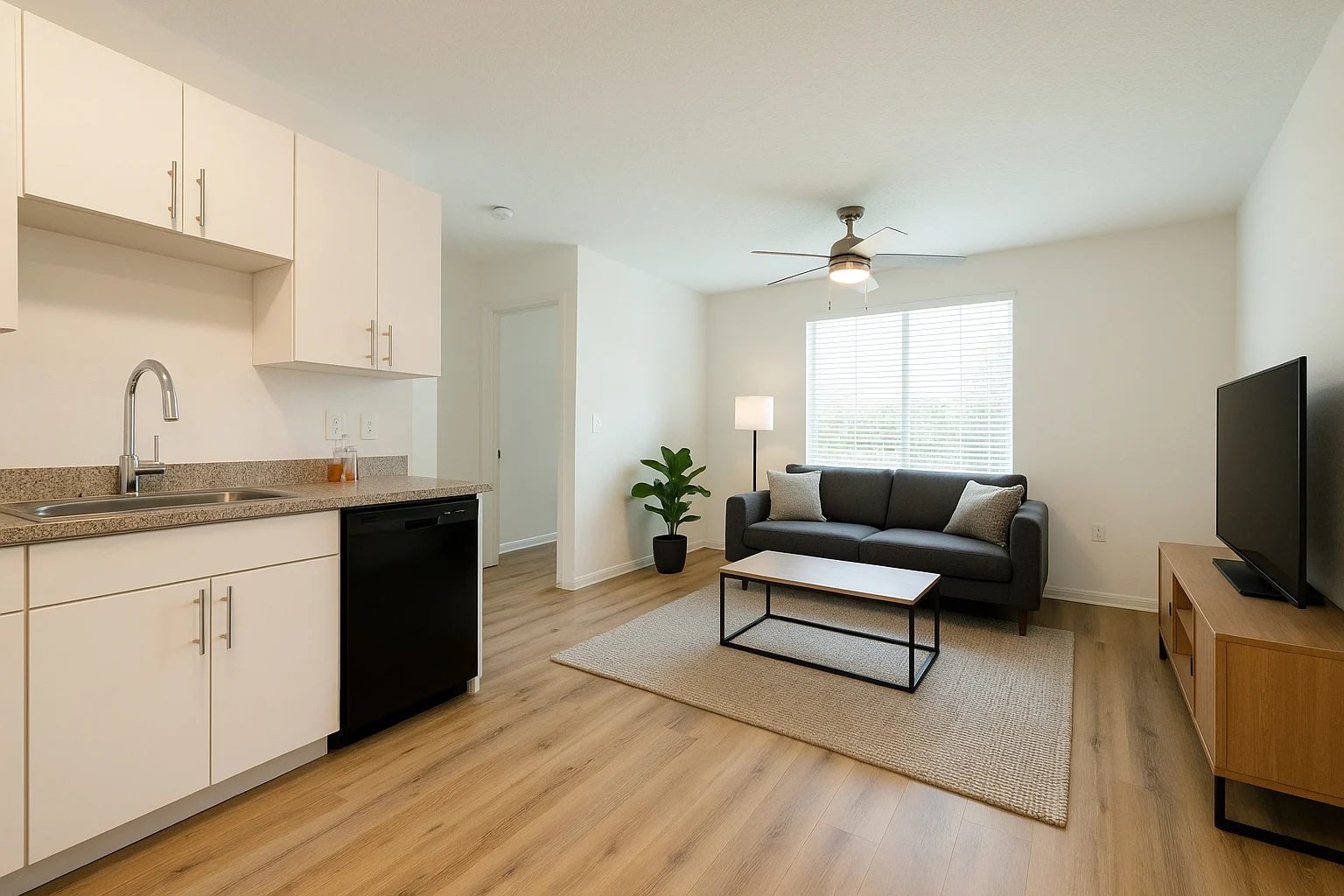 A modern, minimalistic living space featuring a kitchenette with white cabinets and a black dishwasher on the left. A cozy living area is visible on the right, with a dark gray sofa, a coffee table, and a floor lamp. A plant adds a touch of greenery, and natural light streams in through a window with white blinds.