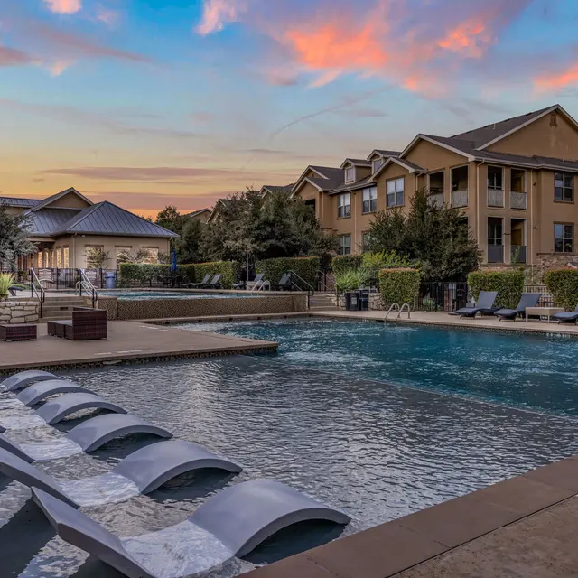 View of a modern apartment complex pool at sunset with lounge chairs and trees in the background.