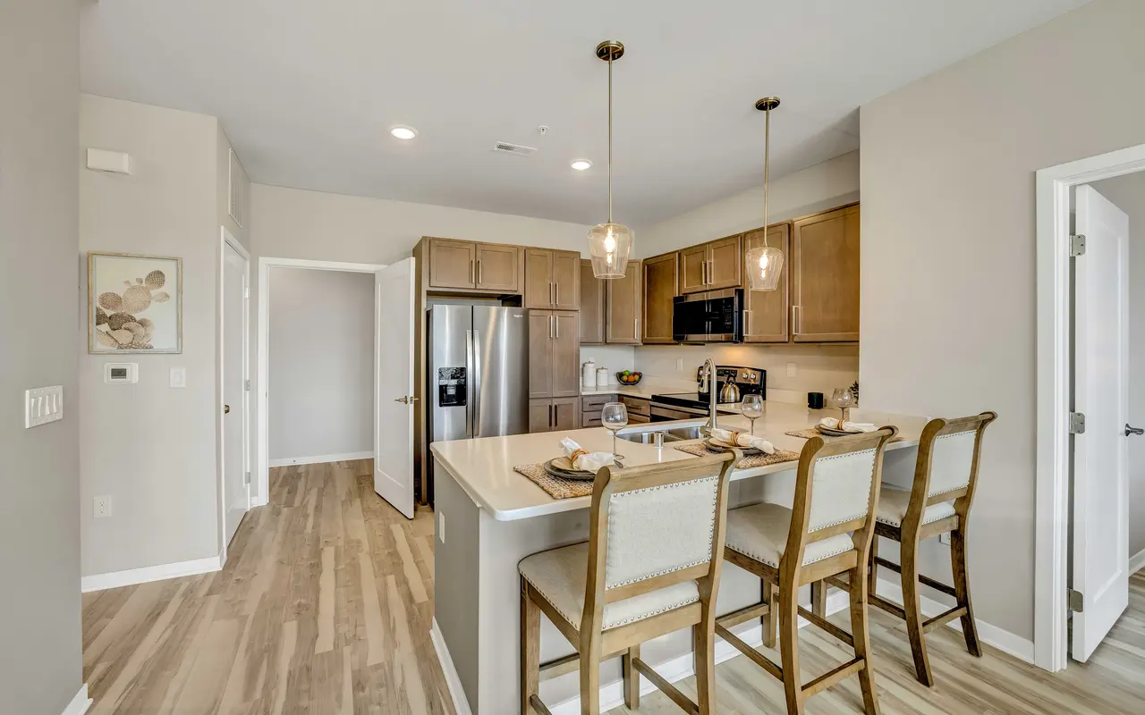 A contemporary kitchen featuring light wood cabinetry, sleek stainless steel appliances, and a spacious countertop with bar stools. Pendant lights hang above the counter, providing illumination.