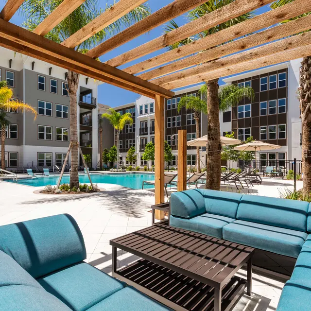 A view of a luxurious outdoor seating area by a pool in an apartment complex, framed by a wooden pergola. The seating features blue cushions and is situated near lush greenery and palm trees, with a swimming pool visible in the background.