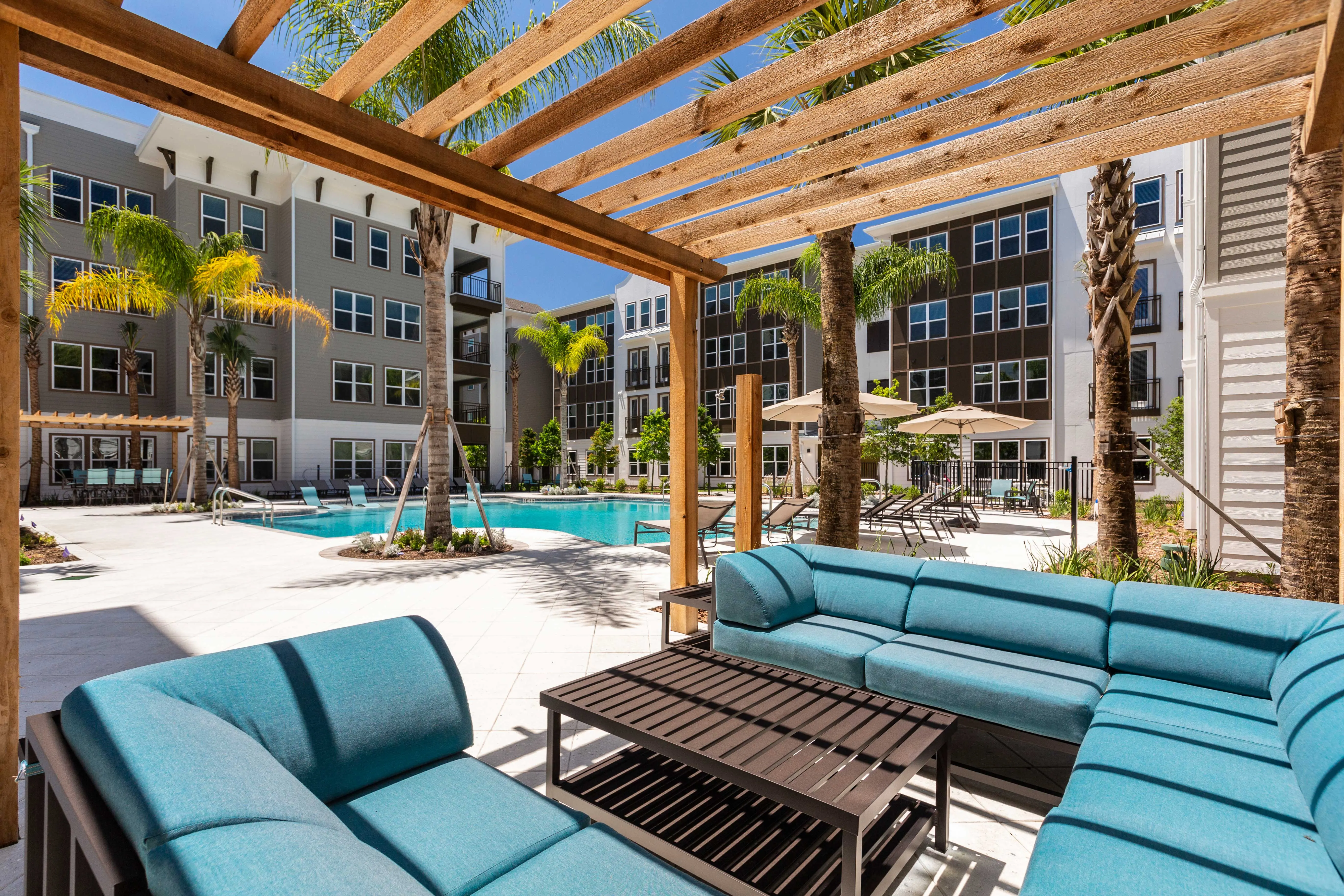 A view of a luxurious outdoor seating area by a pool in an apartment complex, framed by a wooden pergola. The seating features blue cushions and is situated near lush greenery and palm trees, with a swimming pool visible in the background.