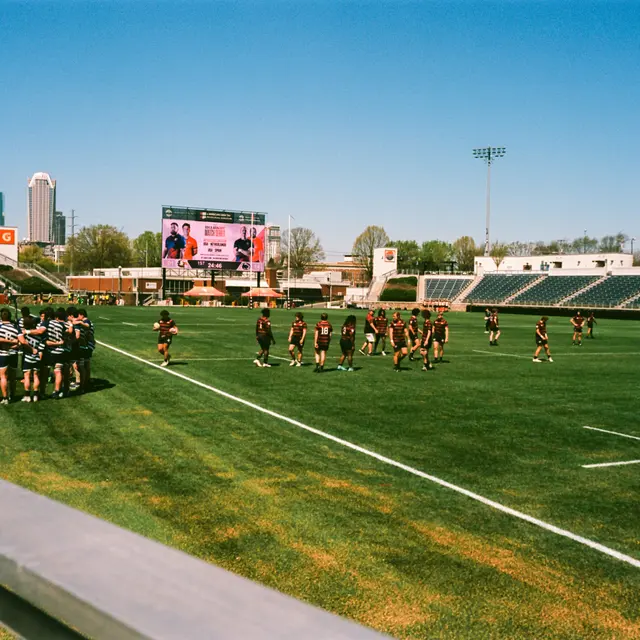 A rugby practice session on a sunny day, with players warming up on the field and a city skyline in the background.