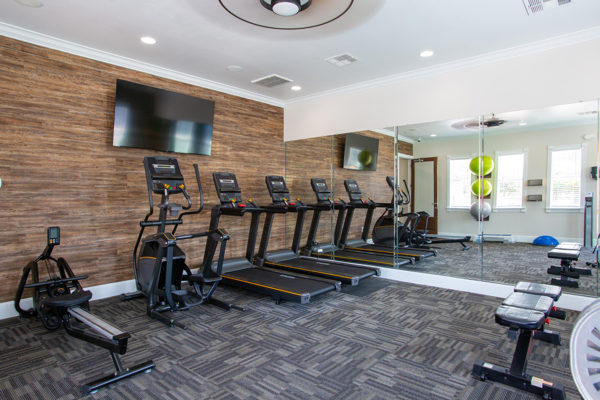 Modern Gym Interior A modern gym interior featuring exercise equipment such as treadmills, a rowing machine, and a weight bench. The walls are adorned with wood paneling and there are large mirrors reflecting the space. In the background, fitness balls can be seen.
