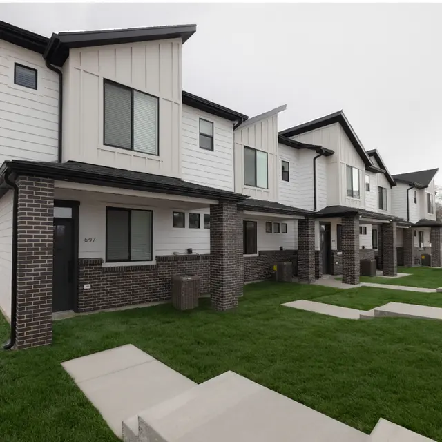 A row of modern two-story townhouses with a mix of white and black exteriors, situated in a neighborhood with green grass and concrete pathways.