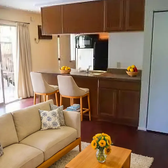 A cozy living room featuring a beige sofa with decorative pillows, a wooden coffee table with flowers, and a kitchenette in the background with bar stools. Natural light streams in from sliding glass doors.