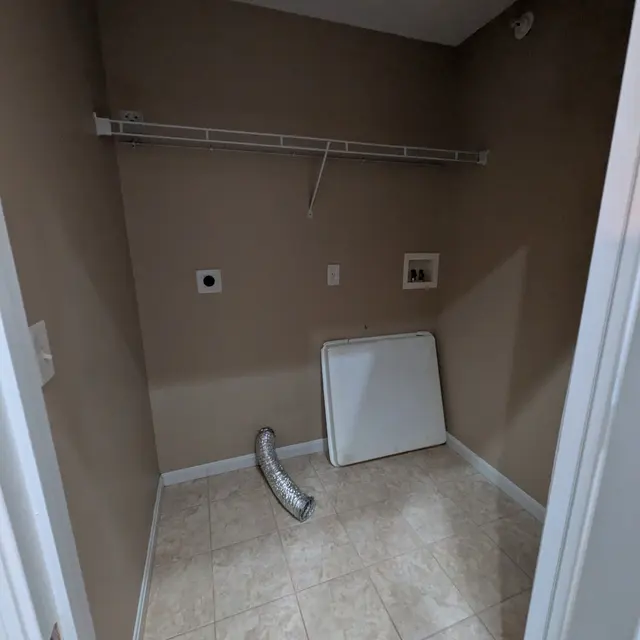An empty utility room with beige walls and tile flooring. There is a wire shelf mounted on the wall, a square white panel on the floor, and a flexible duct lying on the ground.