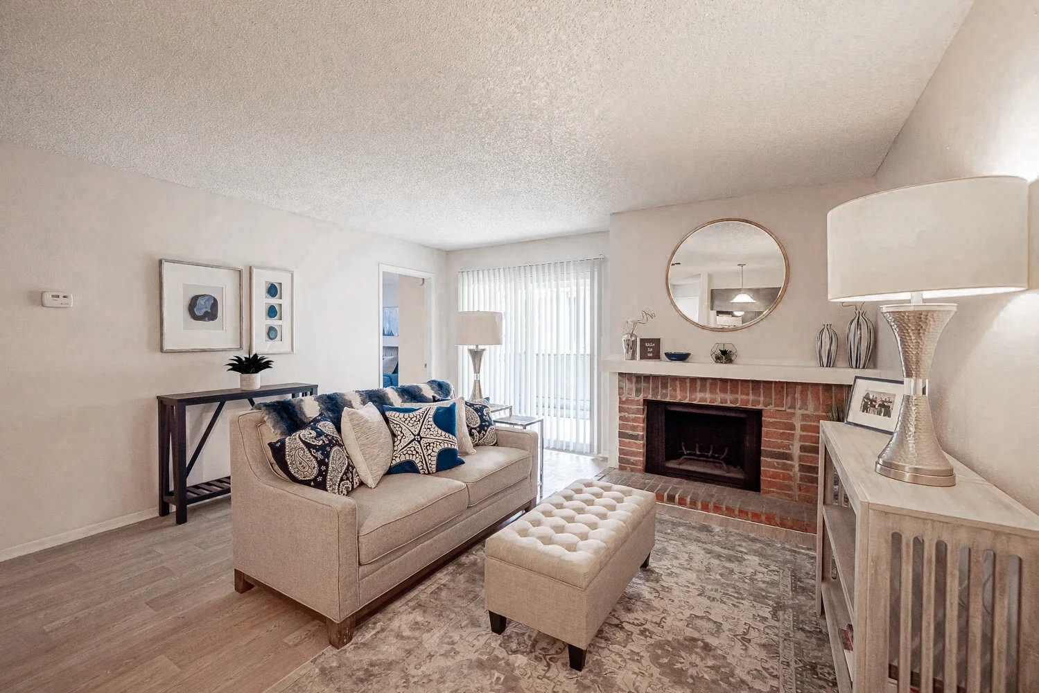 A cozy living room featuring a beige couch adorned with blue and white patterned cushions. There is a small ottoman in front of the couch, a brick fireplace in the background, and large windows with white blinds, allowing natural light to fill the space. Elegant decor items are placed on shelves and tables throughout the room.
