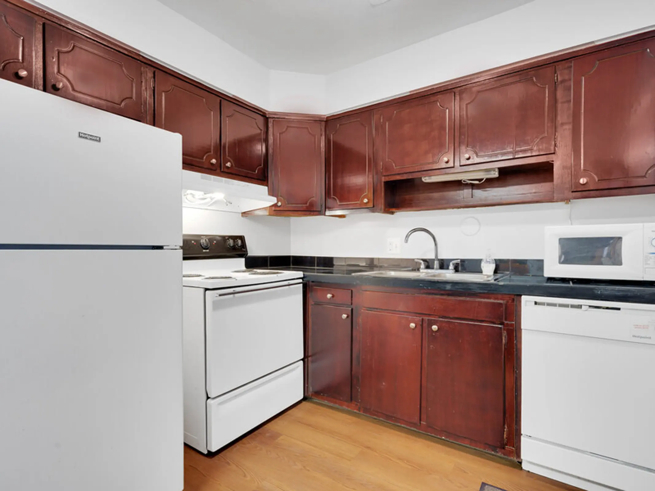A kitchen featuring dark wooden cabinets, a white refrigerator, an oven, and a microwave, with a black countertop and wooden flooring.