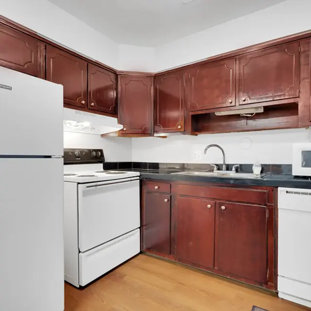 A kitchen featuring dark wooden cabinets, a white refrigerator, an oven, and a microwave, with a black countertop and wooden flooring.