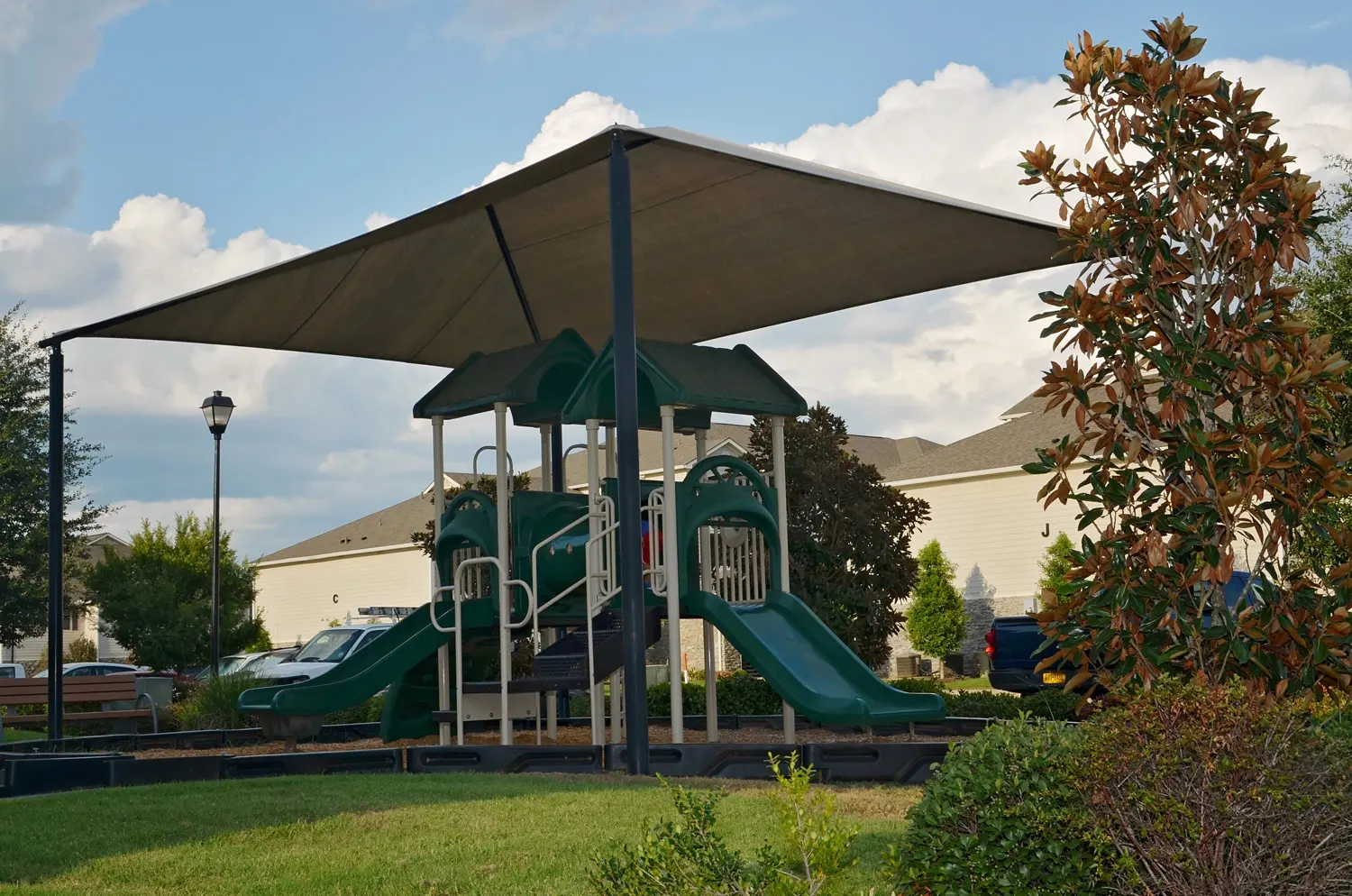 Playground Under Shade Canopy A green playground structure with slides and climbing features under a large shade canopy. Surrounding greenery and a residential background are visible.