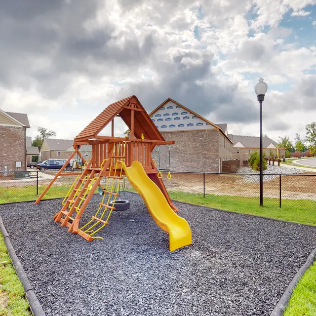 A wooden playground structure with a slide, climbing features, and a tire swing, surrounded by a black rubber surface and a fence. In the background, there are residential houses and a cloudy sky.