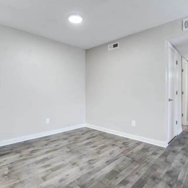 An empty room with light gray walls and wood-like flooring, featuring a single overhead light and a doorway leading to a hallway.
