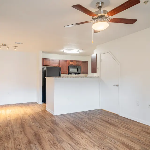 Modern Apartment Living Room Interior view of an empty apartment living room with wooden flooring, a ceiling fan, and a partial kitchen view in the background.