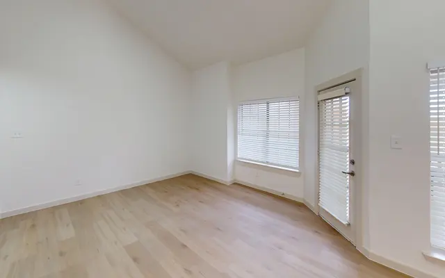 The Gleason An empty living room space with high ceilings and large windows, featuring light hardwood flooring and white walls.