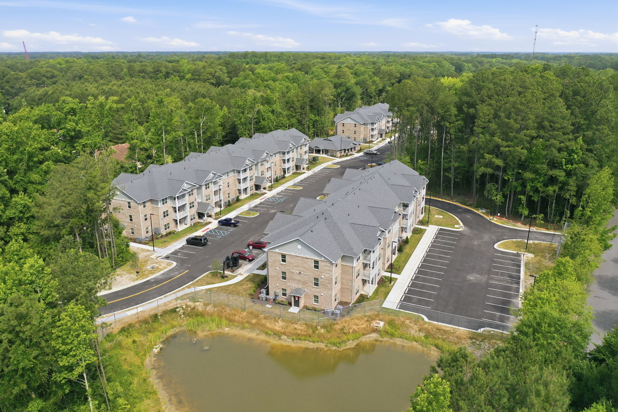 Aerial View of Apartment Complex Aerial view of an apartment complex surrounded by trees and a pond, showing multiple building structures and parking lots.