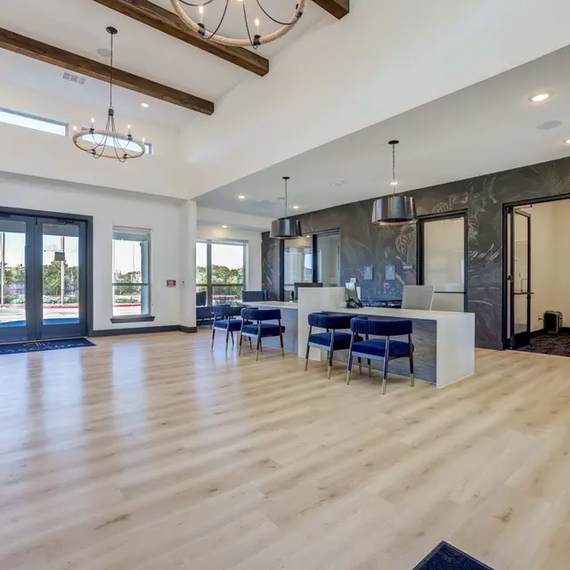 A spacious, modern interior lounge featuring wooden beams on the ceiling, large windows letting in natural light, and a sleek kitchen area with a countertop and bar stools. The room has hardwood flooring and plants in the corners.