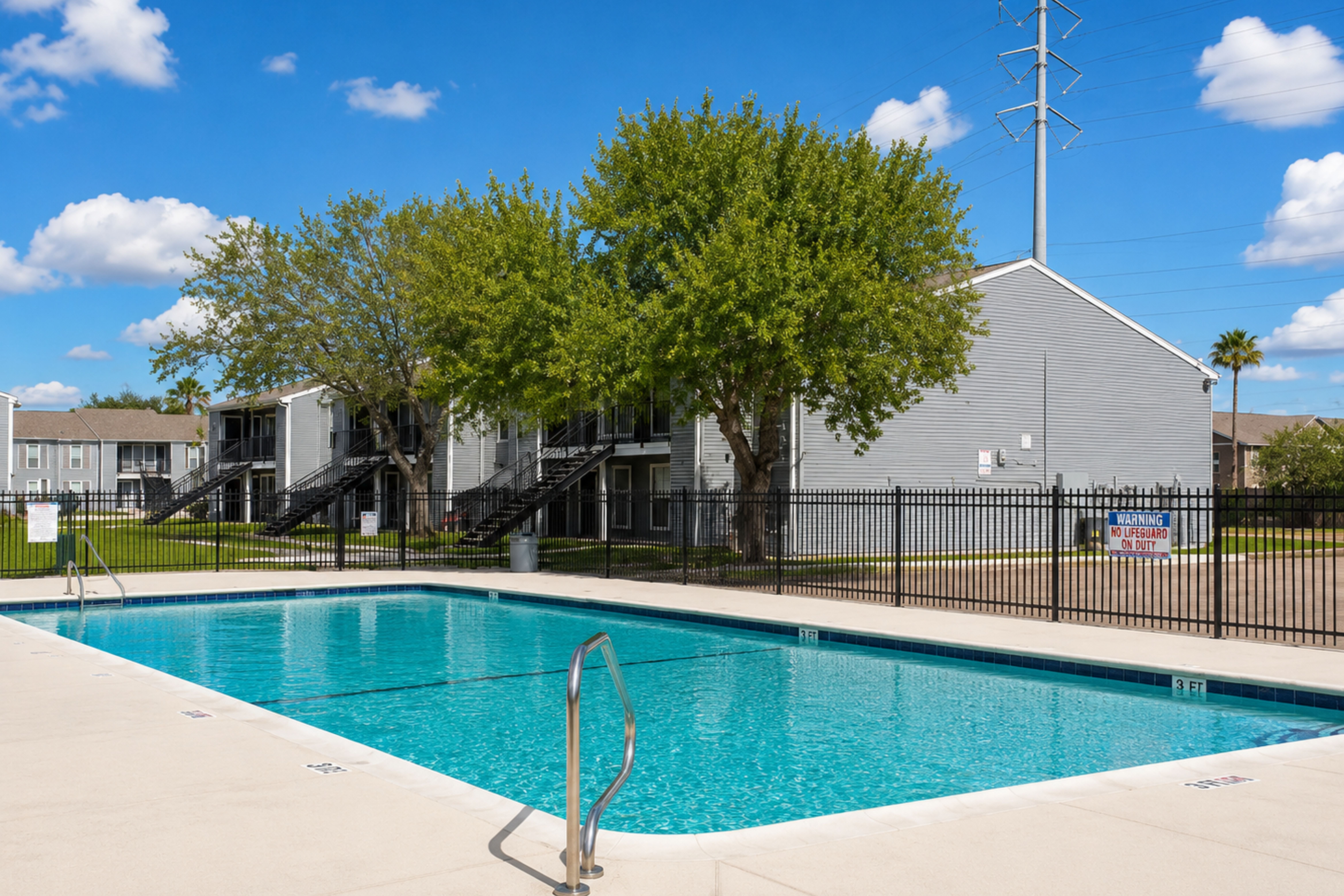 Apartment Swimming Pool Area A view of a swimming pool at an apartment complex, surrounded by green trees and a fence, with buildings in the background under a blue sky with clouds.