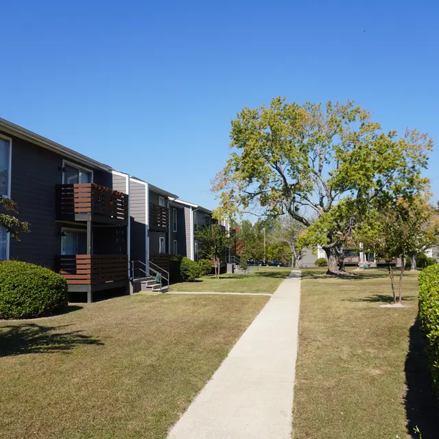 A walkway leading through a landscaped area of an apartment complex, bordered by manicured bushes and trees under a clear blue sky.