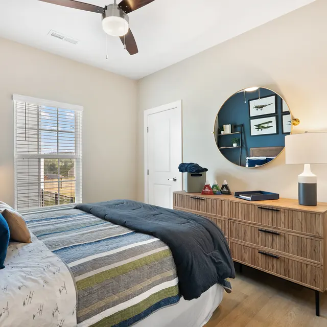 A cozy bedroom featuring a bed with striped bedding, a wooden dresser, and a round mirror on the wall. A bedside lamp illuminates the room, and there's a window providing natural light.