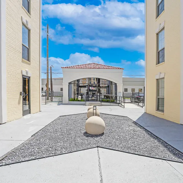 A modern courtyard with a sleek design featuring large round stones on a gravel bed, flanked by two multi-story buildings under a bright blue sky with clouds.