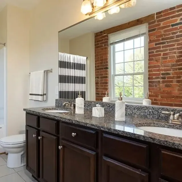 A stylish bathroom featuring a double sink vanity with dark wood cabinets, a granite countertop, a large mirror, and exposed brick walls. A shower area with a striped curtain and a toilet can also be seen.