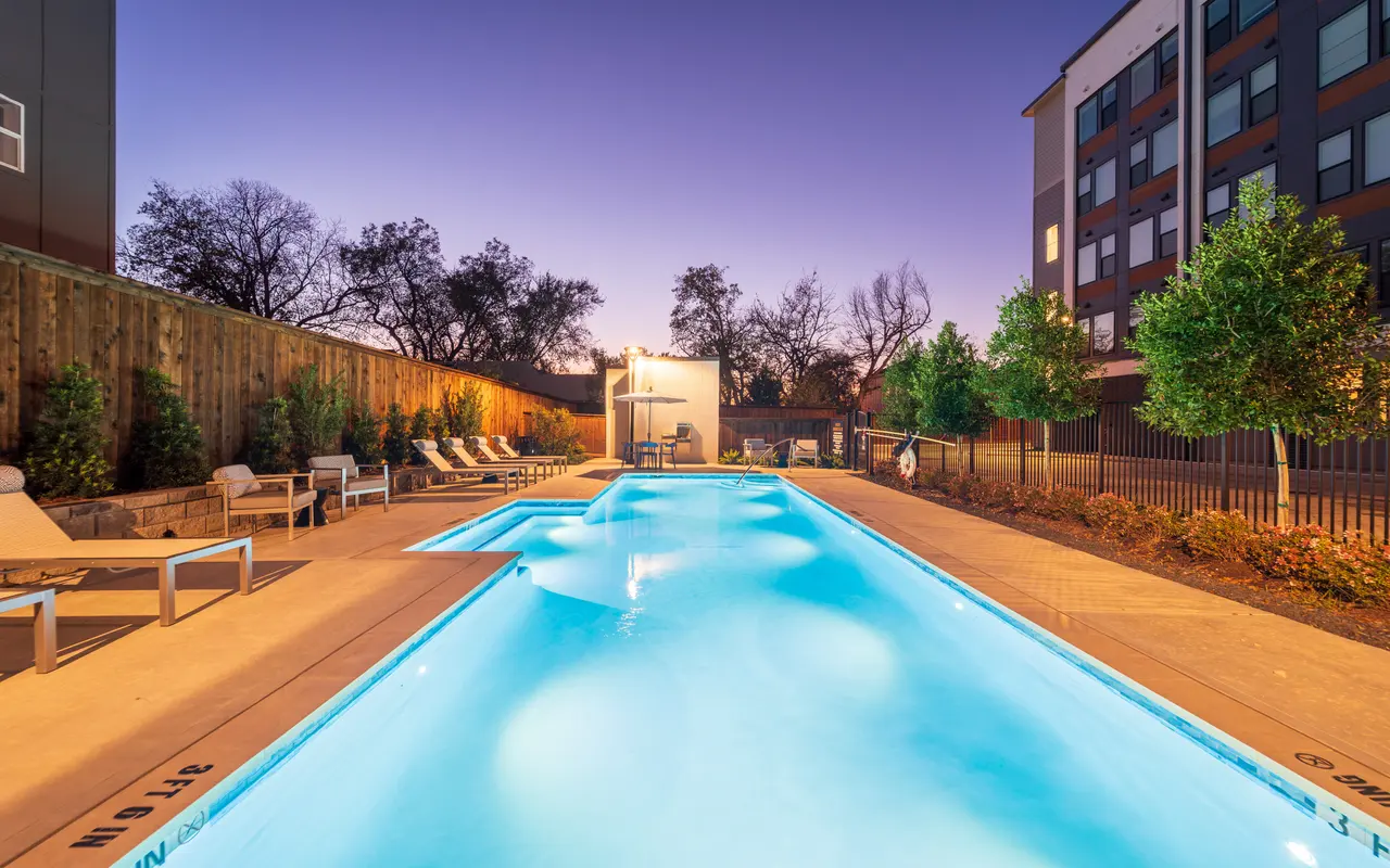 A serene swimming pool area at dusk, featuring a long pool with illuminated edges, lounge chairs on the side, and trees lining the background.