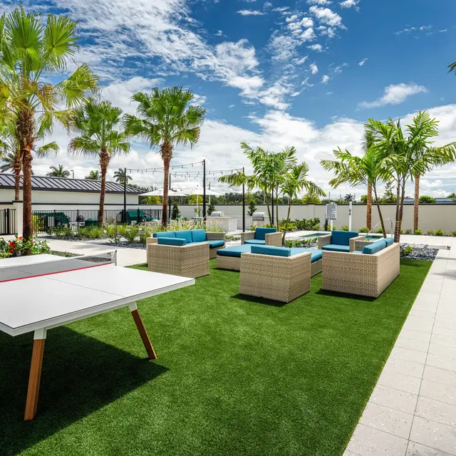 An outdoor living area featuring a ping pong table on green artificial grass, surrounded by palm trees and modern seating arrangements.