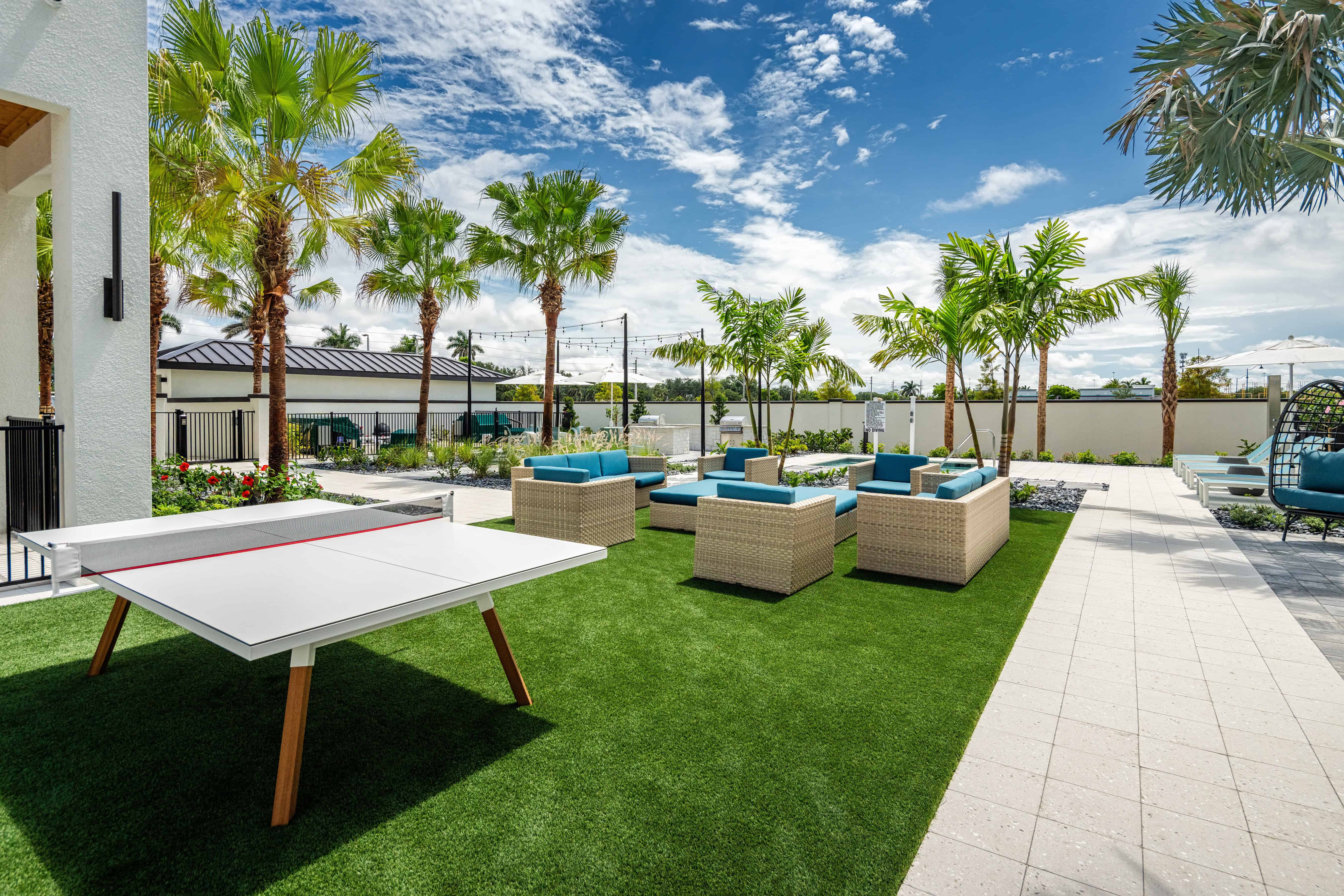 An outdoor living area featuring a ping pong table on green artificial grass, surrounded by palm trees and modern seating arrangements.