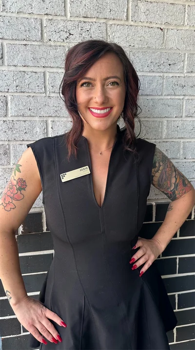 A woman with dark hair dressed in a black outfit, standing confidently with her hands on her hips against a brick wall. She has visible tattoos on her arms and is smiling, wearing a name tag.