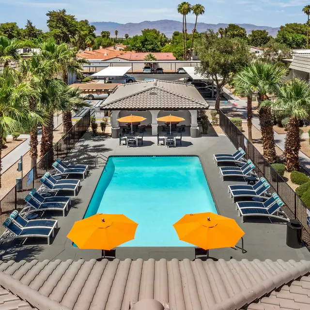 An aerial view of a swimming pool surrounded by lounge chairs and palm trees, with orange umbrellas providing shade. Apartment buildings can be seen in the background, and the setting has a sunny atmosphere.