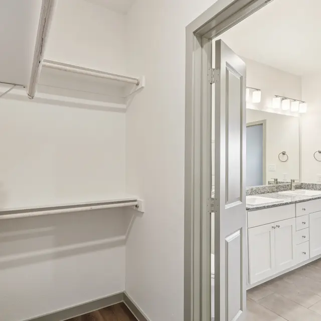 A spacious bathroom area with double sink vanity and a walk-in closet. The bathroom features light-colored countertops, illuminated by overhead lights. The adjoining walk-in closet has a shelf for storage.
