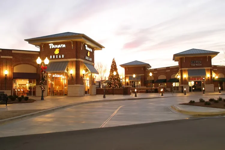 A shopping center featuring a Panera Bread restaurant illuminated at dusk. A Christmas tree is visible in the plaza, enhancing the festive atmosphere with holiday lights.