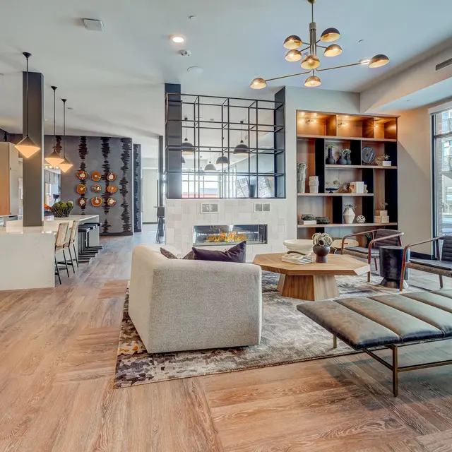 A modern living room featuring a cozy seating area with a light-colored sofa, a lounge chair, and a decorative wooden coffee table. The space is designed with warm wooden flooring and elegant lighting fixtures. A kitchen area can be seen in the background, and large windows provide natural light.